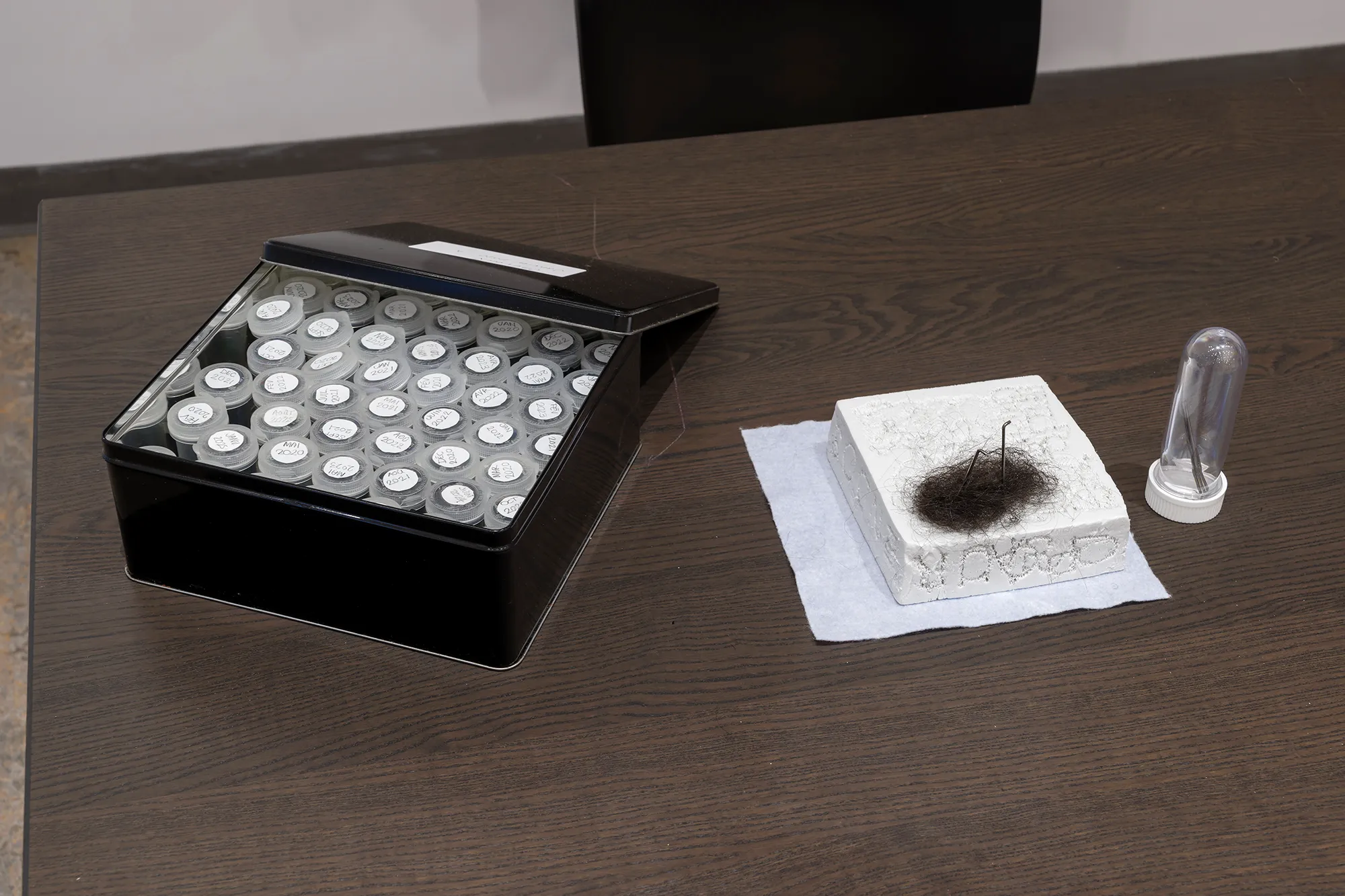 Nadia Gagné, Étiquette, 2023, Performance de longue durée de feutrage à l’aiguille sur nappe de coton blanc tissé, cheveux, fil d’acrylique, latte de bois et aimants. (Accessoires)